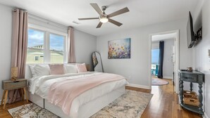 Ein Schlafzimmer mit einem Bett, einem Deckenventilator, einem Gemälde, einem Nachttisch, einer Kommode und Blick ins Freie.