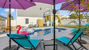 Ein Garten mit Pool, aufblasbaren Flamingos und Liegestühlen unter violetten Sonnenschirmen.
