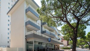 Un hôtel moderne à plusieurs étages, doté d’une façade en verre, de nombreux balcons et d’un arbre au premier plan.