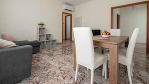 Un coin repas avec une table en bois et des chaises blanches, un canapé gris et une petite étagère contenant des livres et une plante.
