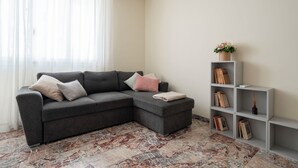 Un salon avec un canapé d’angle gris, un tapis blanc et une étagère grise contenant des livres et un vase avec des fleurs.