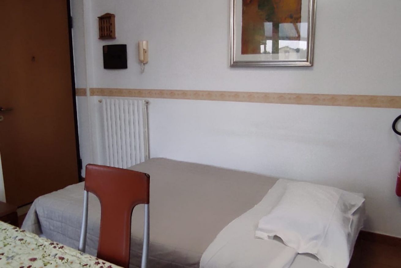 A single bed with a white comforter and pillows, a wooden chair, a radiator, a wall-mounted phone, and a framed picture.