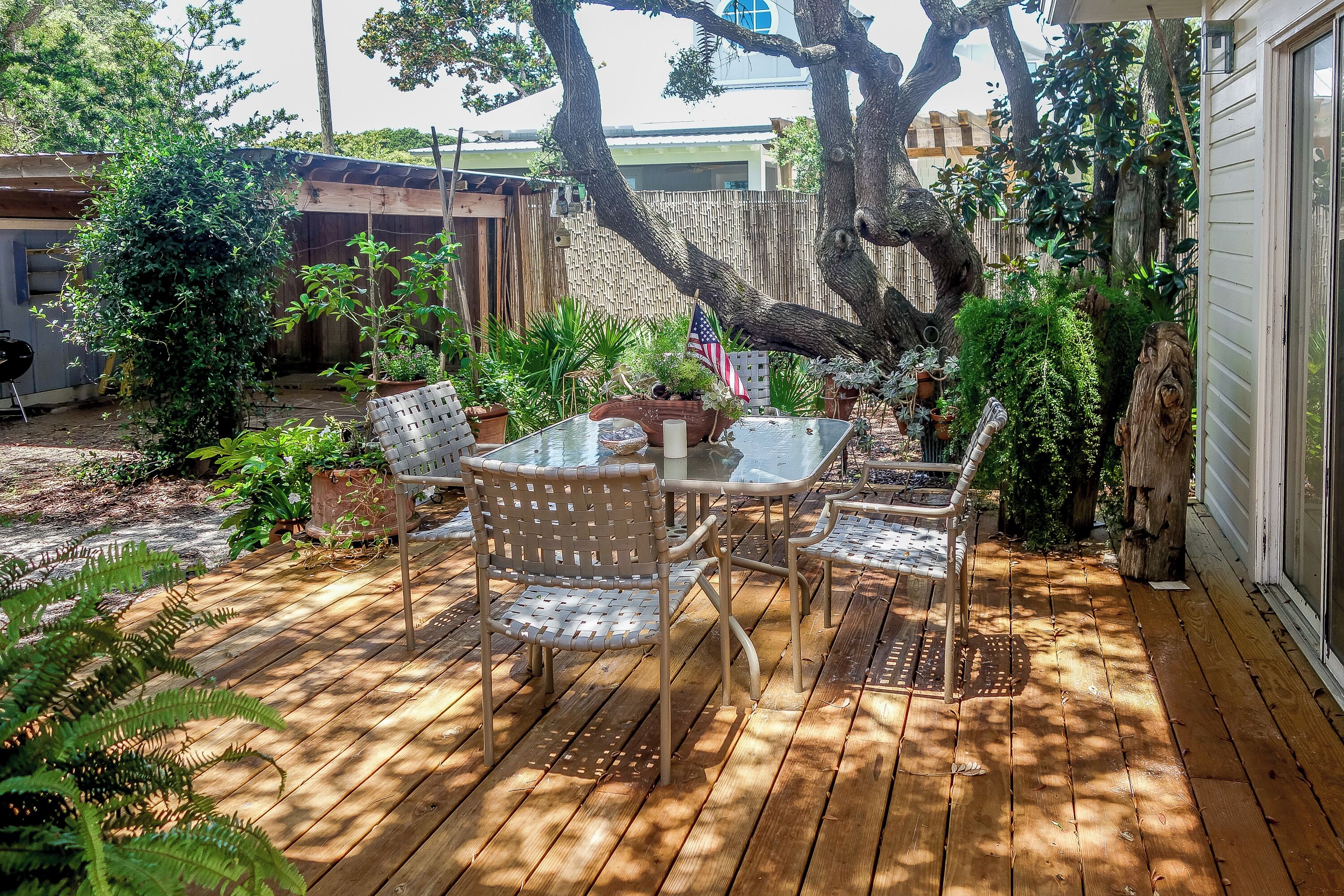 A wooden deck with a glass table and chairs, surrounded by potted plants and a large tree.