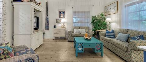 A cozy living room with a television, a patterned sofa, a teal coffee table, and a wicker chair.