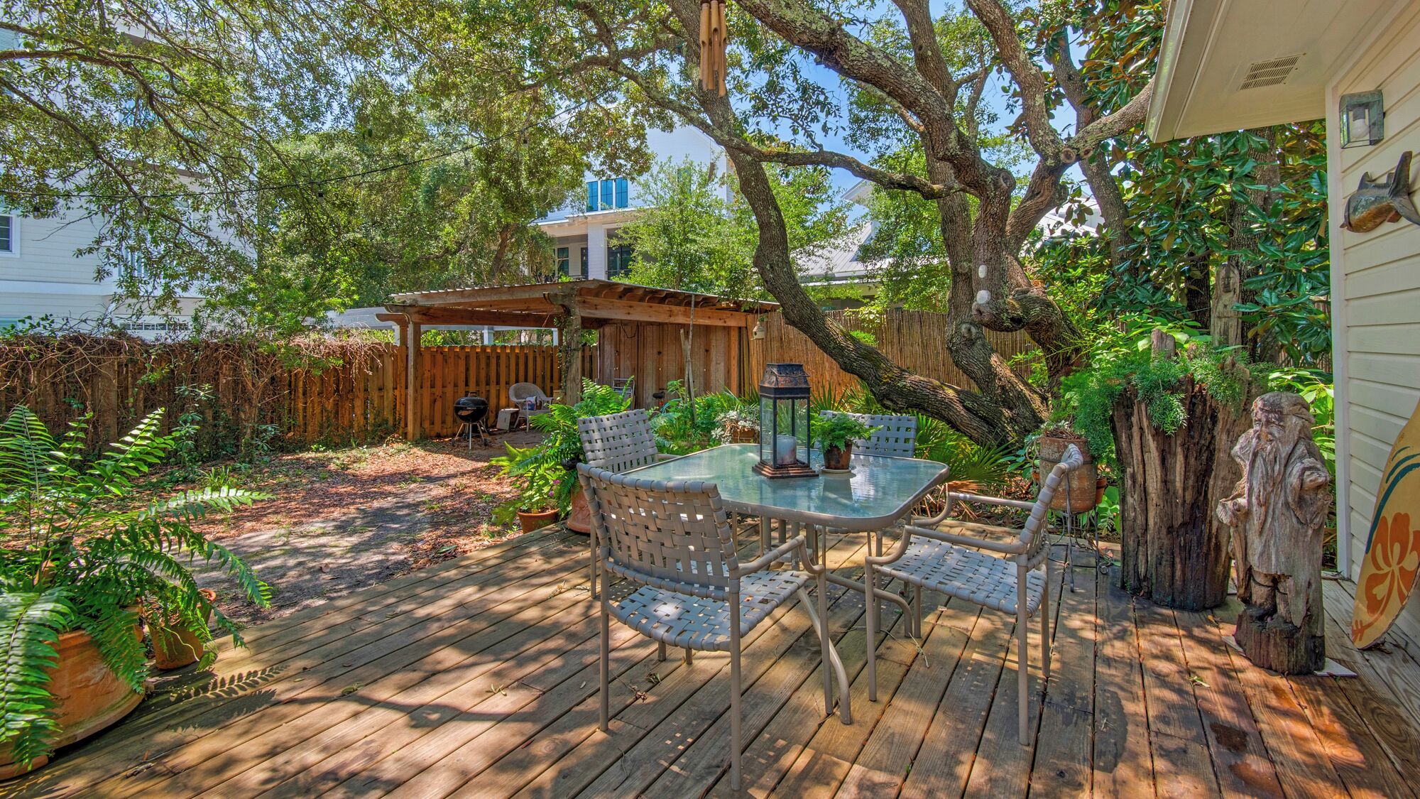 A wooden deck with a glass table and chairs, surrounded by lush greenery and a wooden pergola.