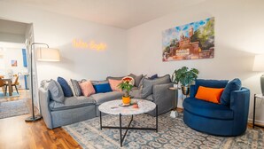 Un salon avec un canapé gris, un fauteuil bleu, une table basse ronde et un tableau représentant un bâtiment accroché au mur.