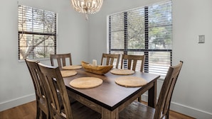 A dining room with a wooden table and chairs, a chandelier, and large windows with blinds.