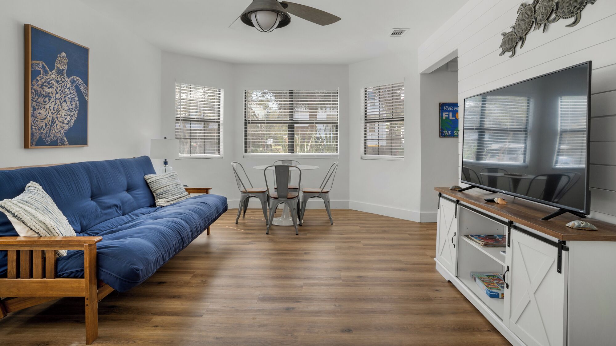 A living room with a blue sofa, a dining table with chairs, a TV on a cabinet, and a ceiling fan.