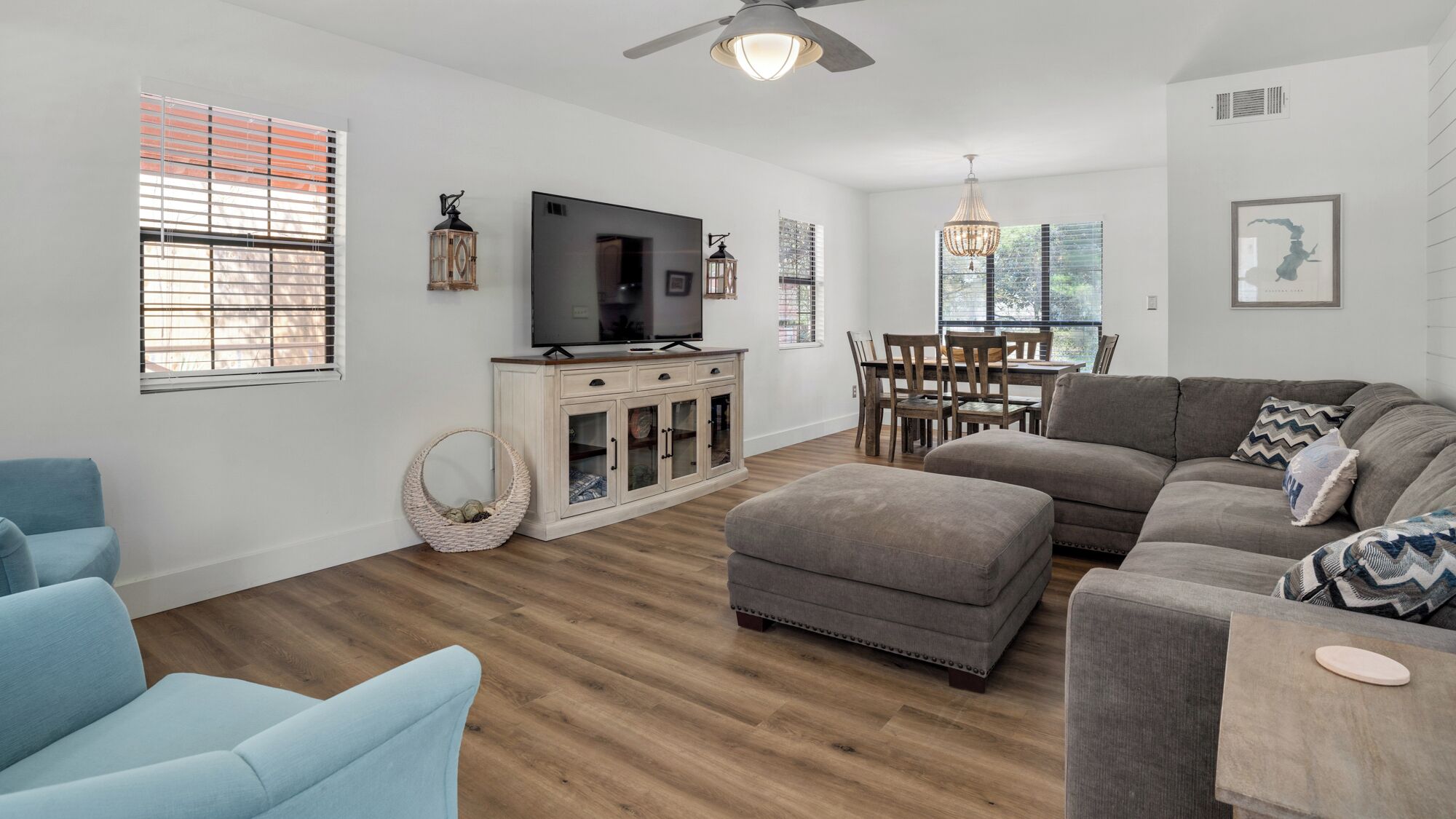 Un salon moderne avec un canapé gris, un fauteuil bleu et un pouf, un plancher en bois et un téléviseur posé sur un meuble.