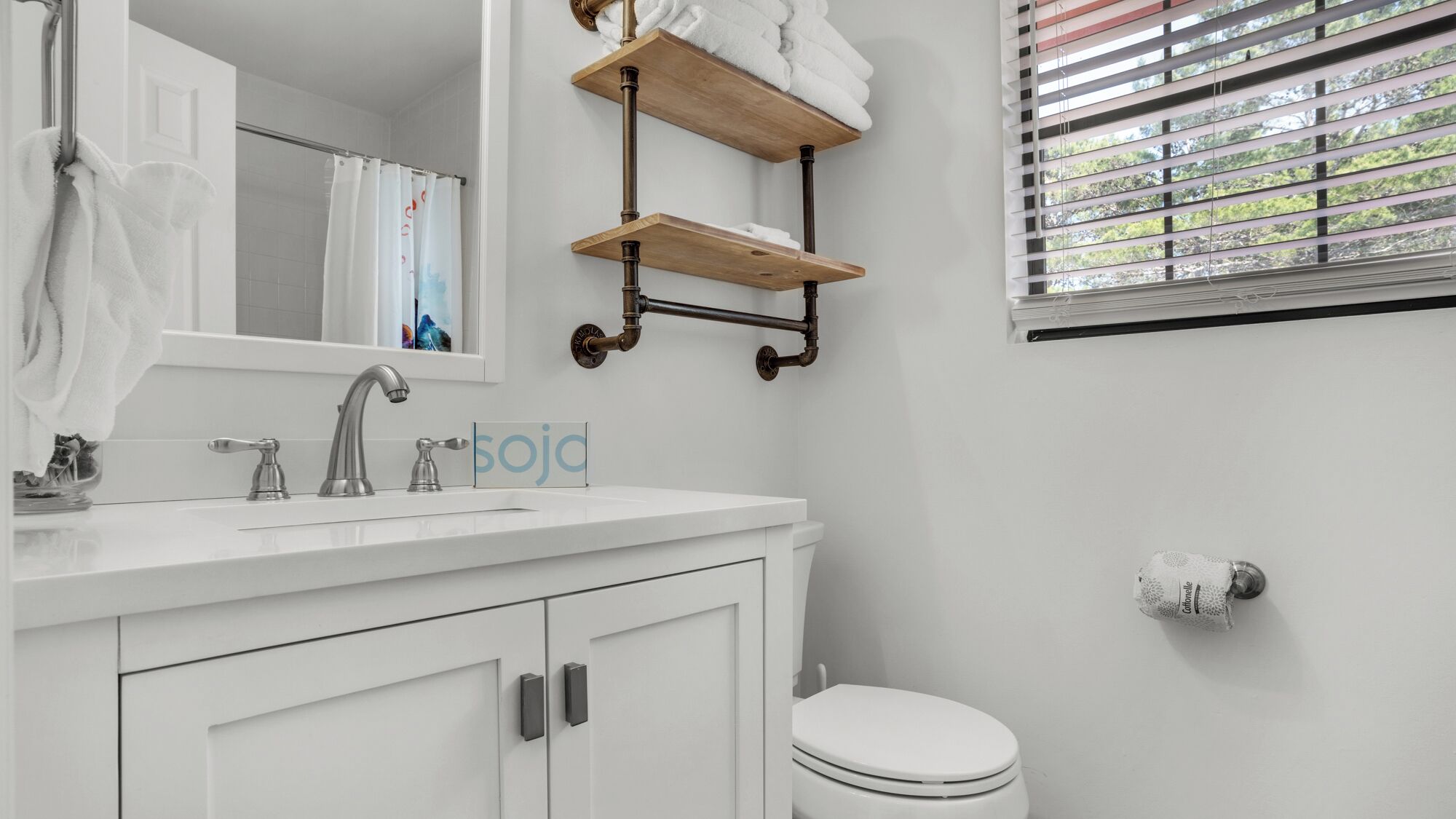 Een badkamer met een witte wastafel, een spiegel, een handdoekrek, een toilet en een raam met zonwering.
