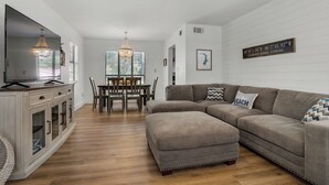 A modern living room with a sectional sofa, a dining table with chairs, a TV, and a chandelier.
