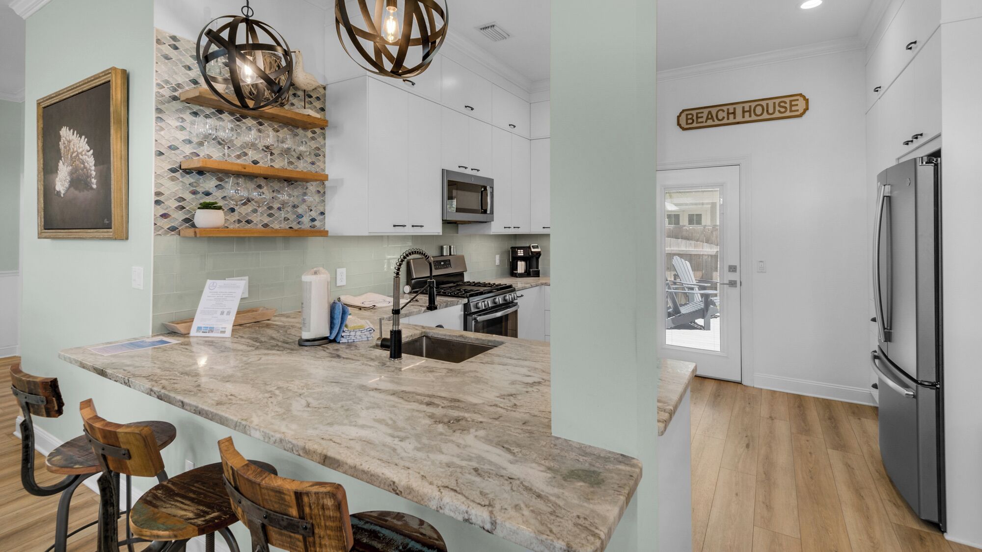 A modern kitchen with a marble countertop, wooden chairs, a built-in oven, and a sign reading "BEACH HOUSE".