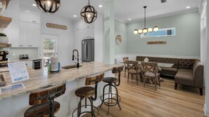 Una cocina con encimera de mármol, taburetes de madera y una zona de bar. Se ve un refrigerador, un lavamanos y una zona de comedor con sofá y sillas.
