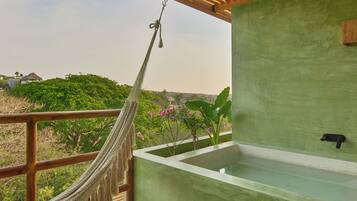 A hammock hanging on a wooden railing, a green wall, and a view of greenery and buildings in the background.