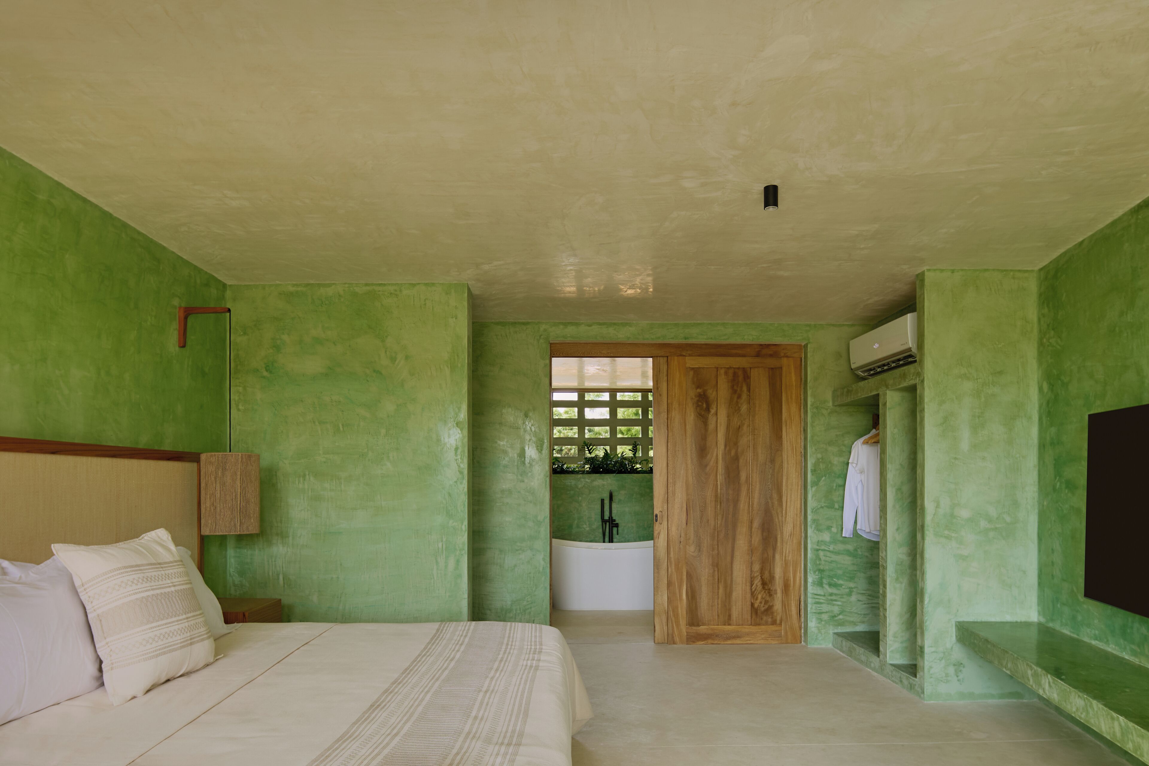 A bedroom with a bed, a wooden headboard, a nightstand, a wall-mounted lamp, a green wall, a wooden door, a bathtub, and a towel rack.