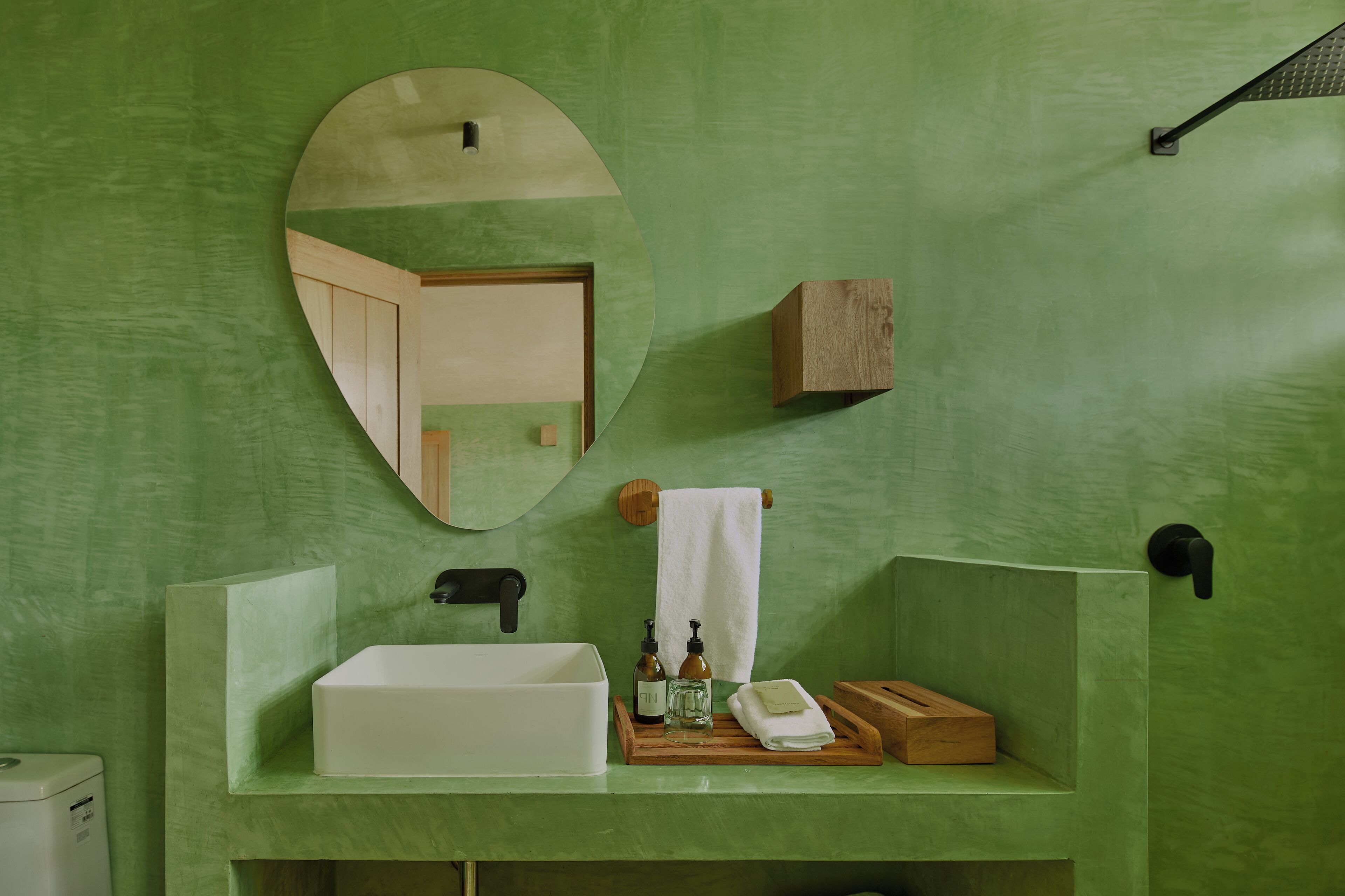Une salle de bain avec un mur vert, un miroir rond, un lavabo blanc et une étagère en bois où sont rangés les articles de toilette.