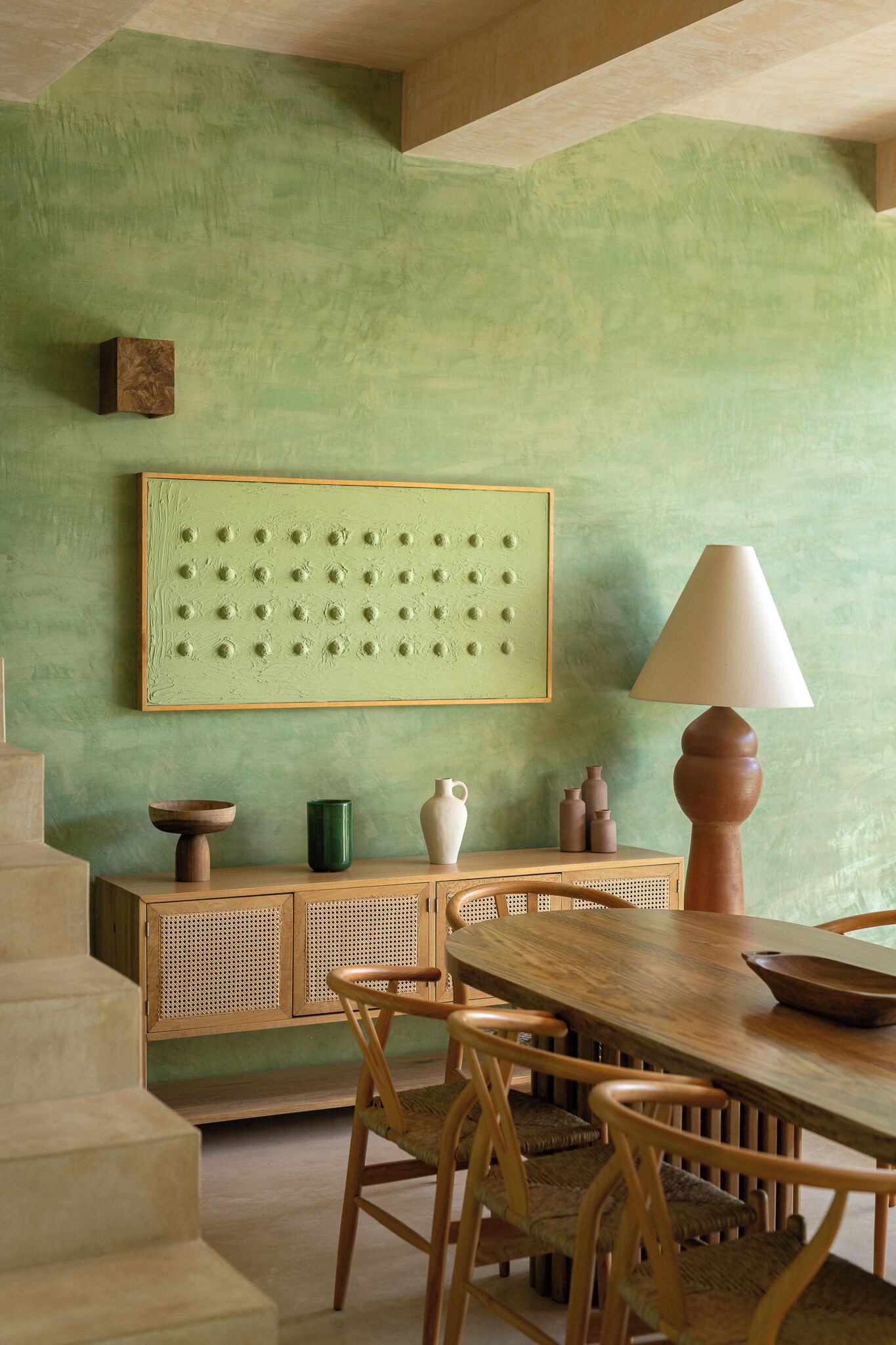 Un espace repas comprenant une table en bois, des chaises et un mur vert agrémenté d’une œuvre d’art à la texture particulière.