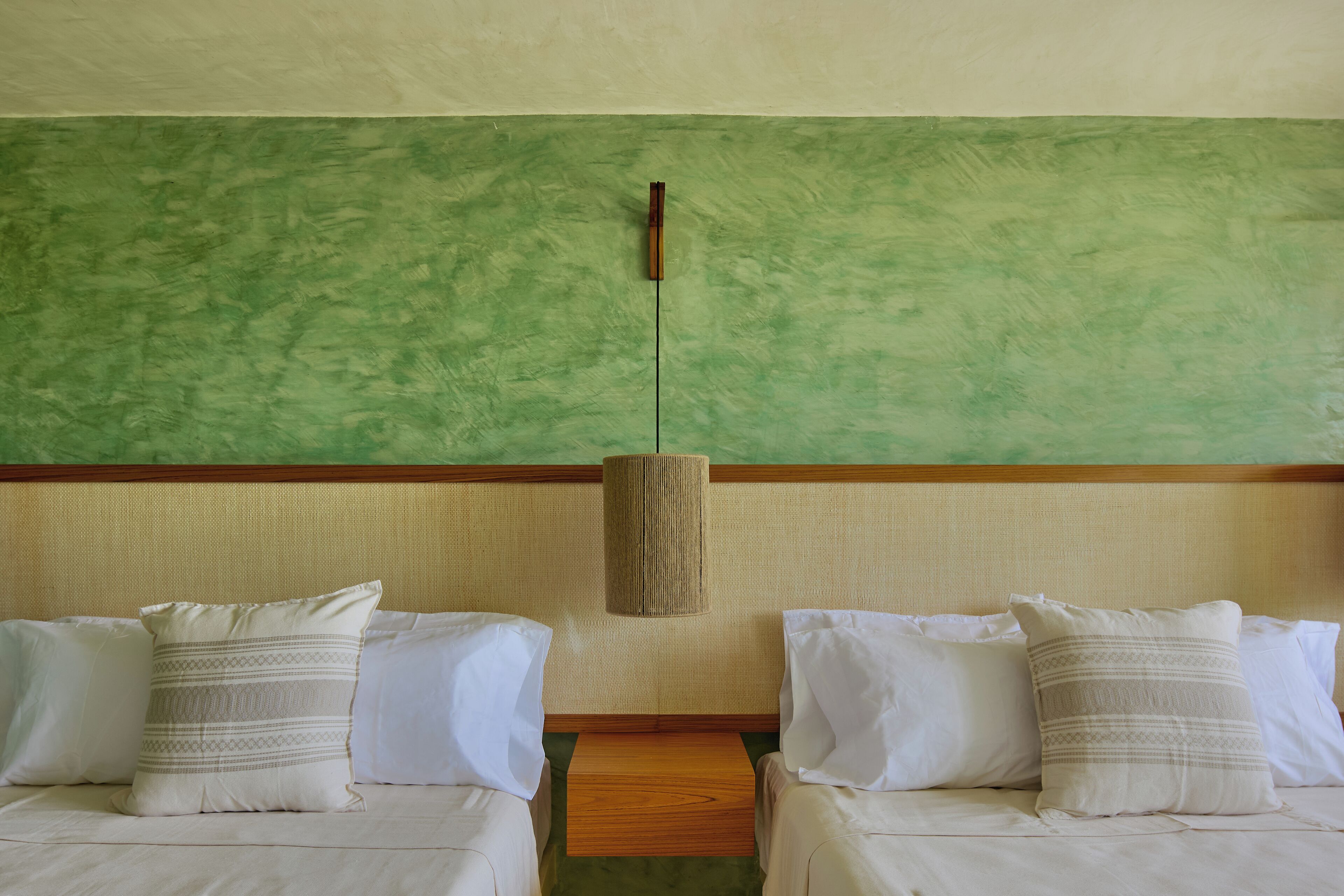 Habitación de hotel con dos camas, cada una con ropa de cama blanca y almohadas estampadas. Una lámpara de techo tejida cuelga del techo, y las paredes están pintadas de verde.
