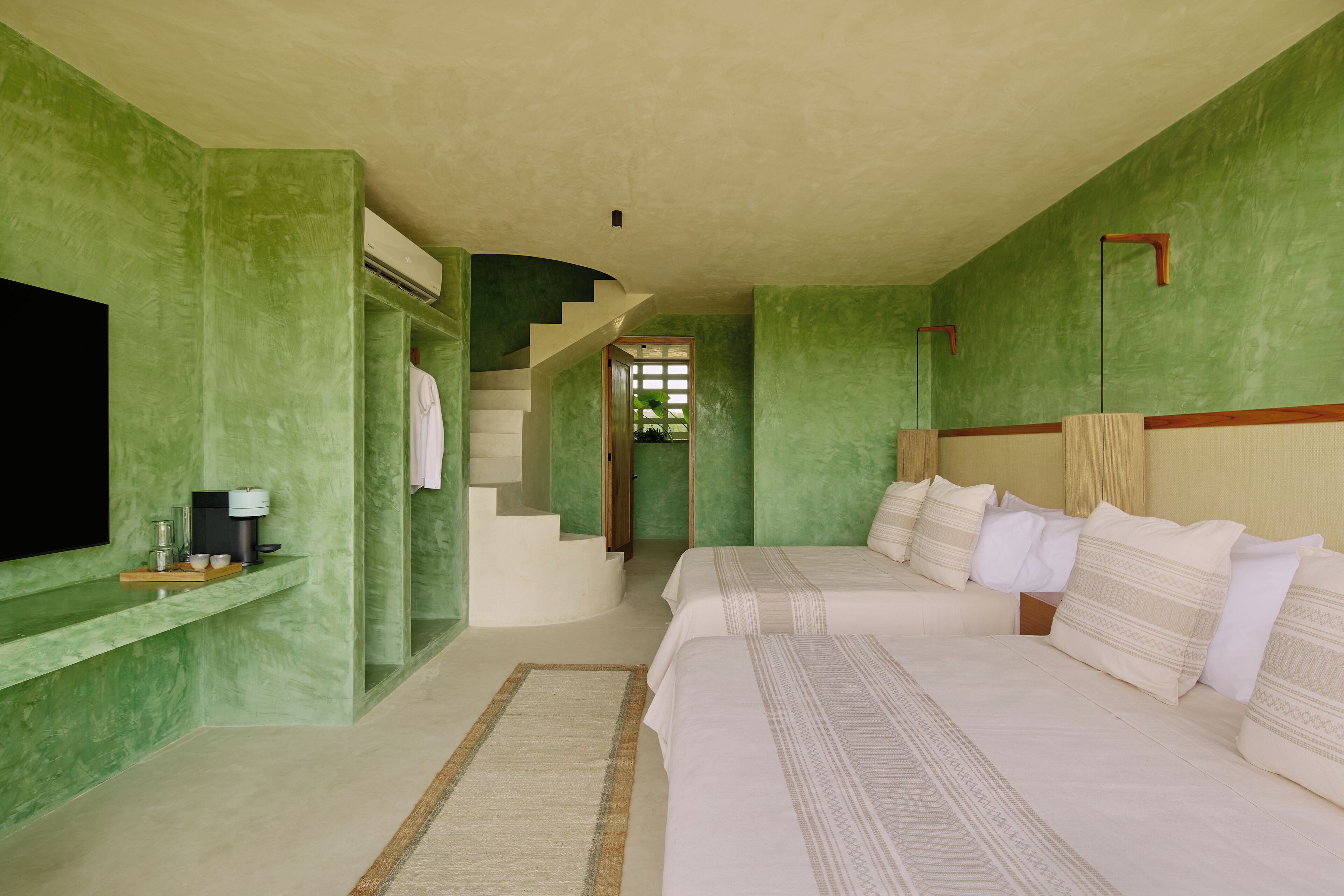 Un dormitorio con pared verde, televisor, mesita de noche, cama con ropa blanca y una escalera que lleva a otra habitación.