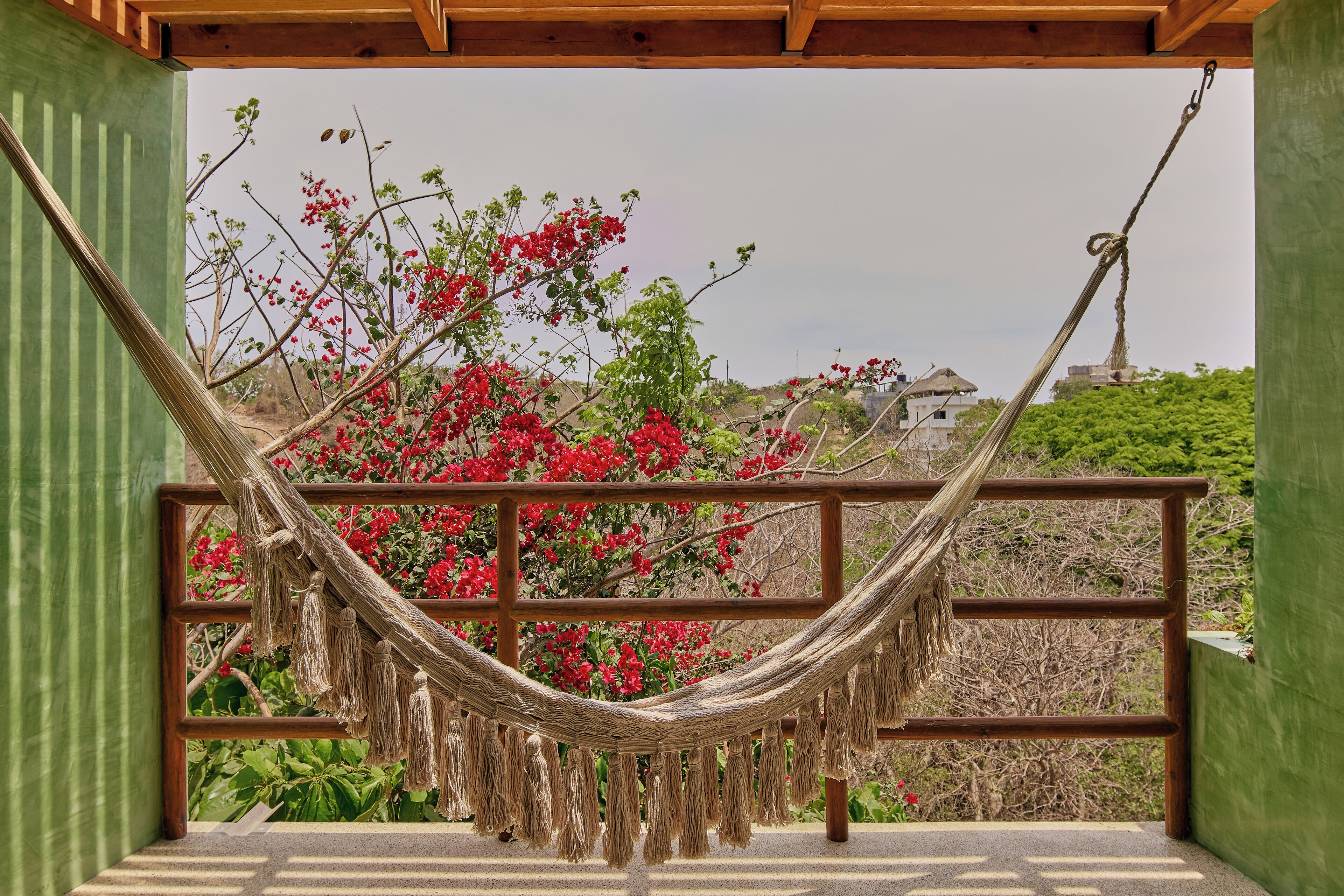 A hammock suspended between wooden beams, with a view of red flowers and a building in the background.