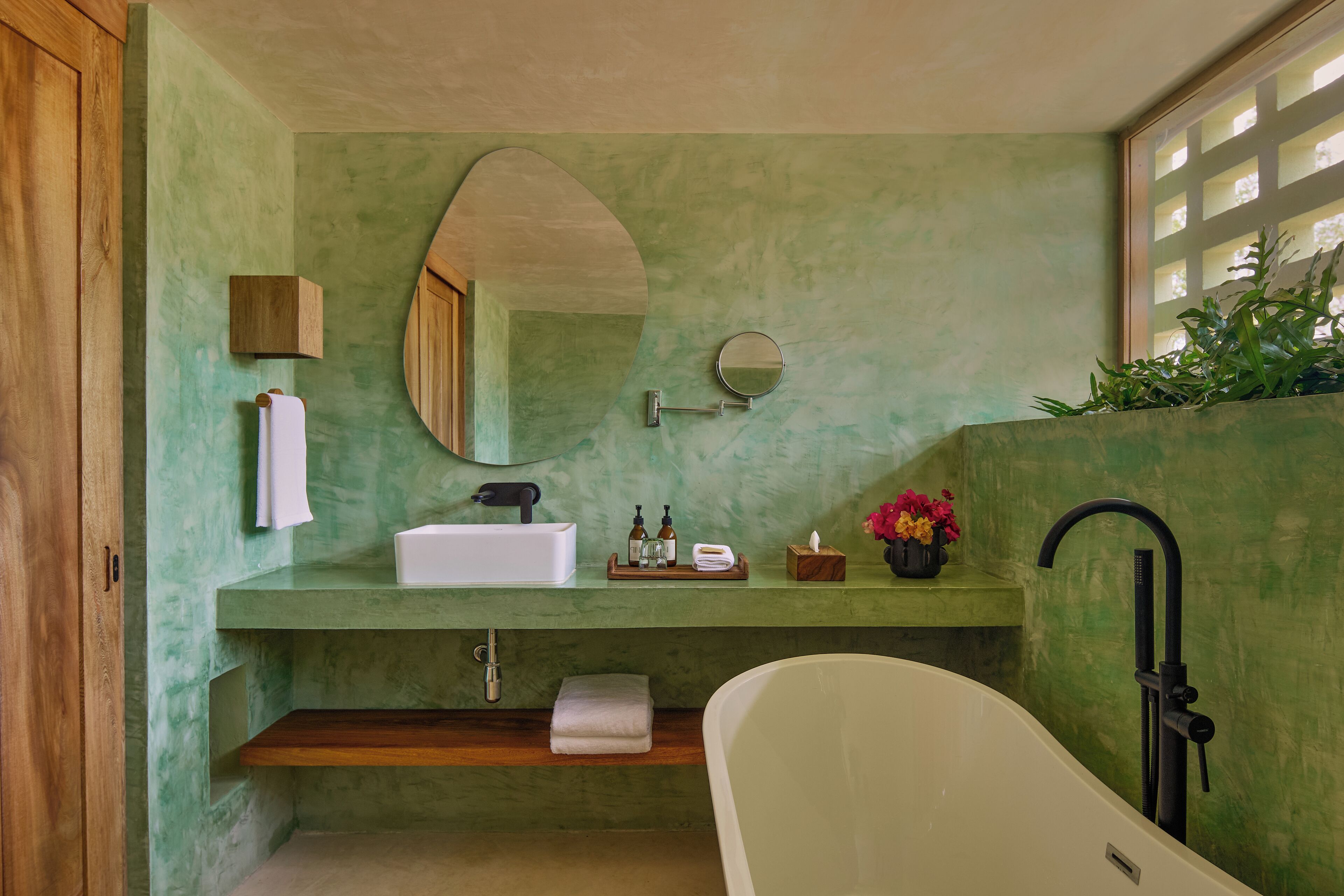 Un baño con una pared verde, una bañera blanca, un estante de madera, un espejo y una ventana con vistas a zonas verdes.