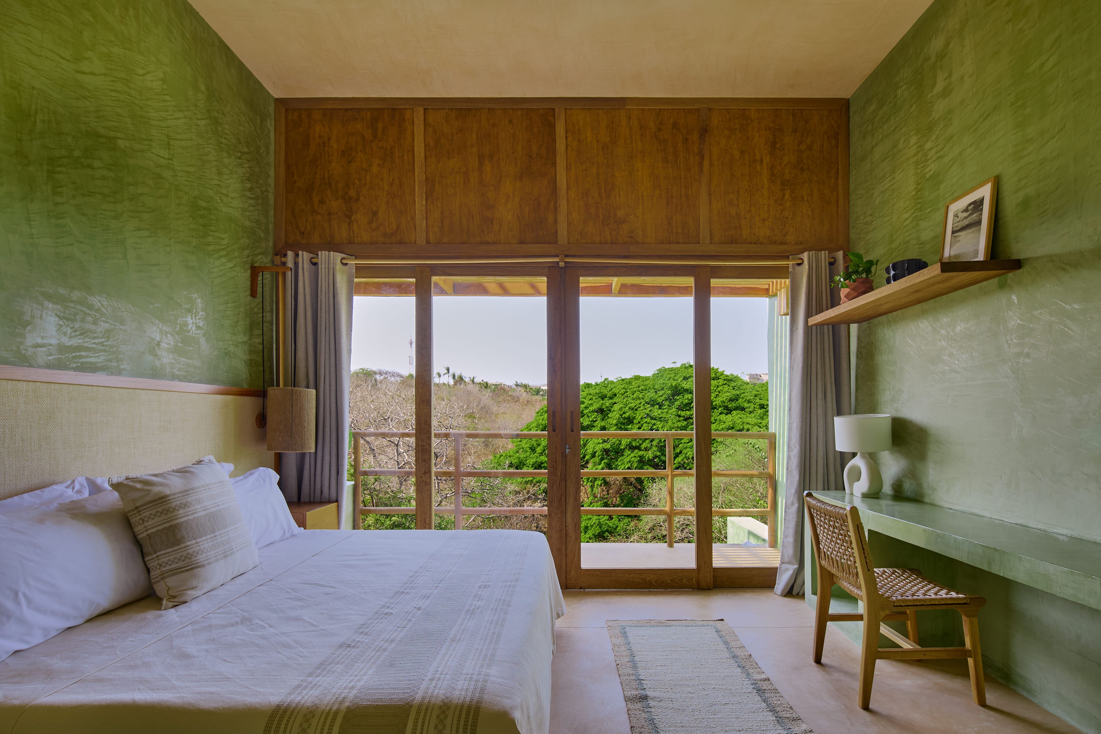 A bedroom with a bed, a chair, a nightstand, a lamp, and a view of greenery through a sliding door.