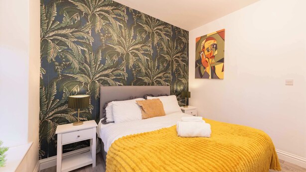 A bedroom with a bed, a nightstand, a lamp, a painting, and a tropical wallpaper.