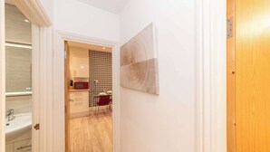 A hallway with a wooden door, a white wall with a textured artwork, a houndstooth patterned wall, and a kitchen area with a microwave.