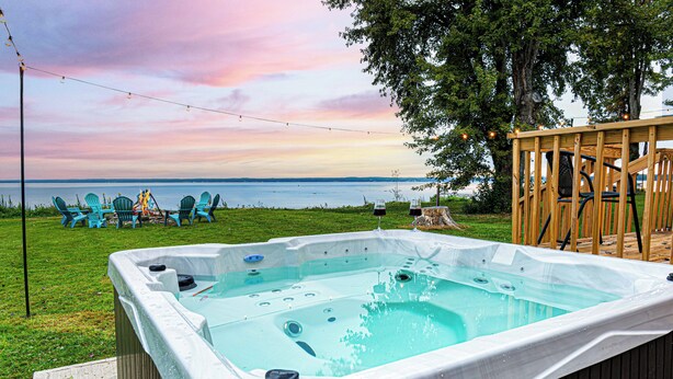 A hot tub with a wooden deck, surrounded by chairs and a view of the water. - Lakefront Modern home. Gorgeous Sunsets! near Sylvan Beach (Canastota)