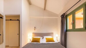 Un dormitorio con una cama, dos lámparas de noche, una mesita de noche, una ventana con cortinas y una puerta que da a otra habitación.