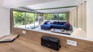 Una sala de estar moderna con un sofá seccional azul, una mesa de centro de madera y un tostador negro. Amplias ventanas ofrecen vistas a zonas verdes al exterior.