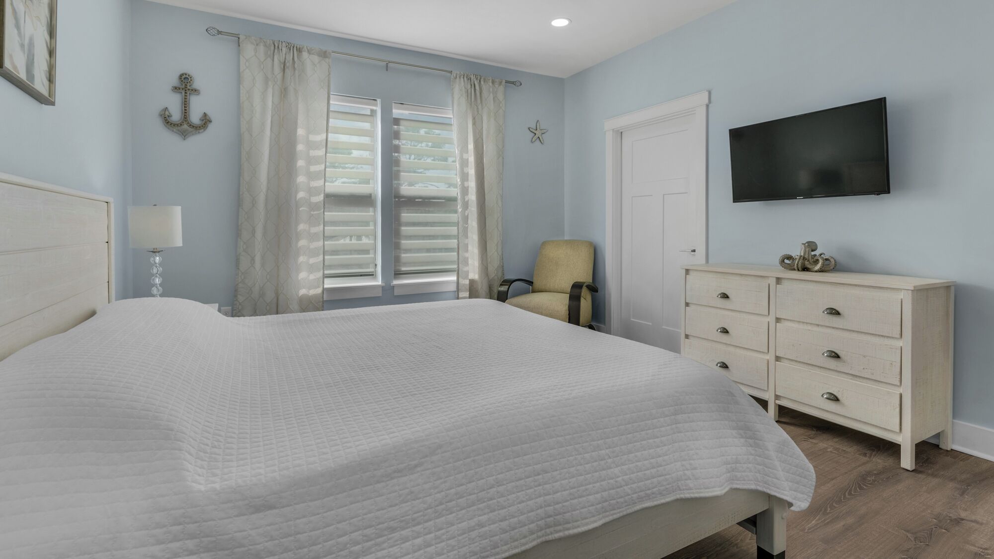 Une chambre à coucher avec un grand lit, une chaise, une commode, un téléviseur et une fenêtre avec des rideaux.