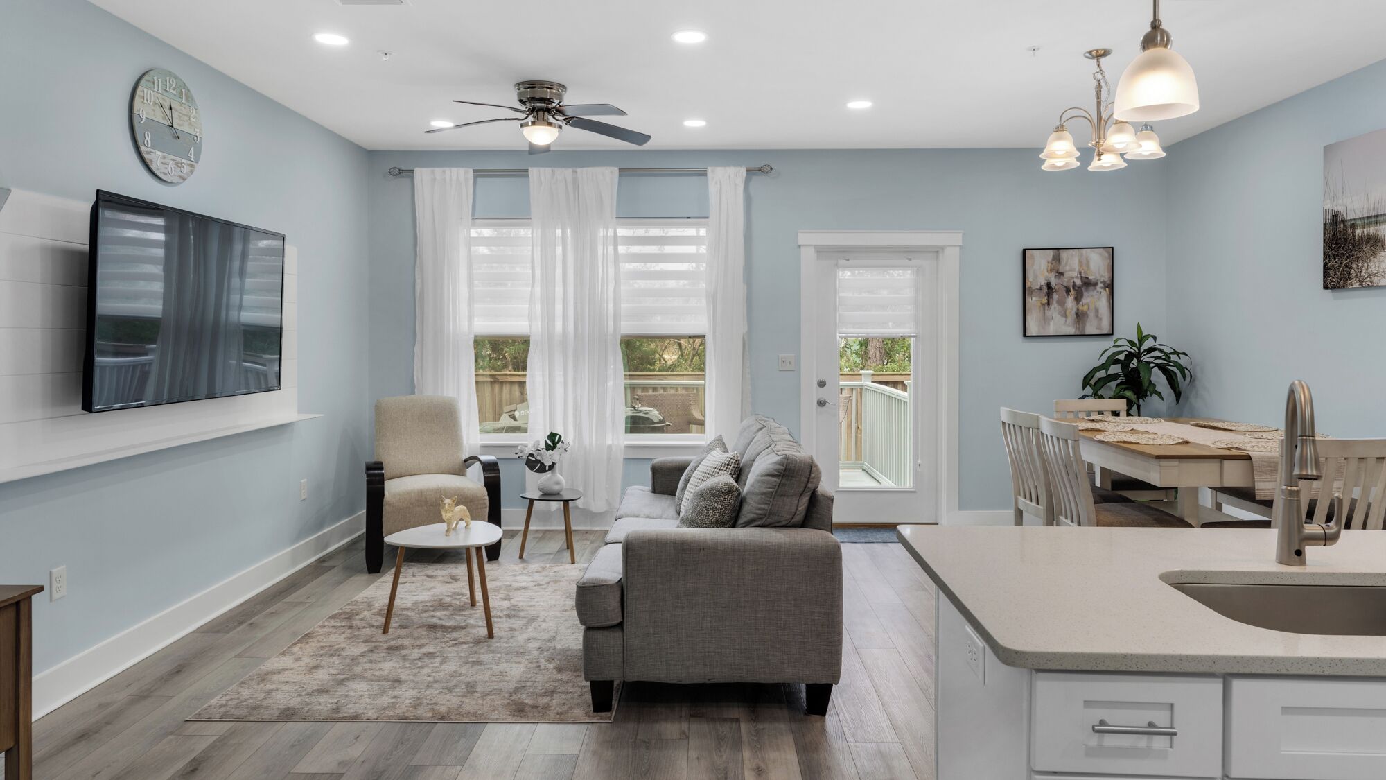 A modern kitchen with a dining area, a ceiling fan, and a large window with white curtains.