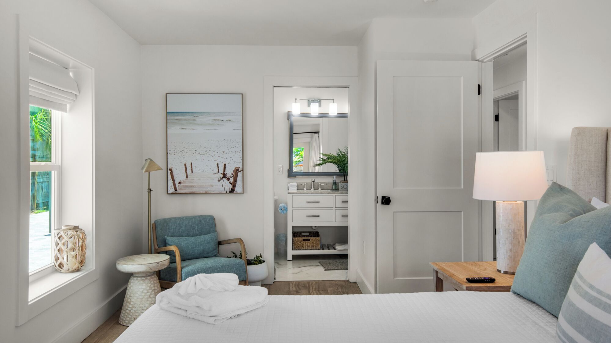 A bedroom with a bed, a chair, a nightstand, a lamp, a painting, and a bathroom visible through an open door.