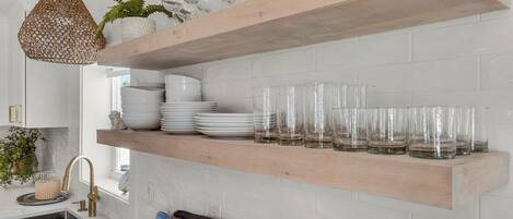 A kitchen with white tiled walls, a wooden shelf holding plates and glasses, a hanging light fixture, and a window with plants.