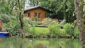 A wooden cabin surrounded by lush greenery and a calm pond with two boats. - Captivating 1-bed Cabin in Newmarket (Newmarket)
