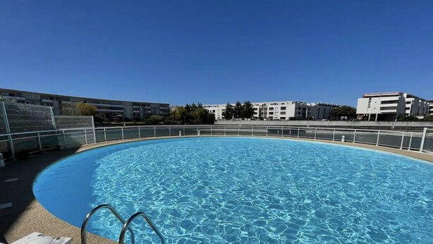 A clear, circular swimming pool with a metal safety railing, surrounded by a low wall and a clear blue sky.