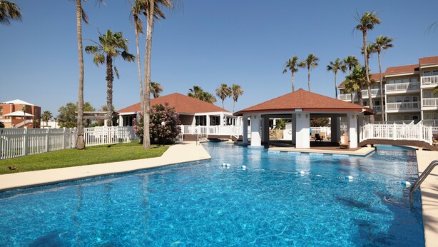 A swimming pool with palm trees and a covered area, surrounded by a white fence and buildings with balconies.