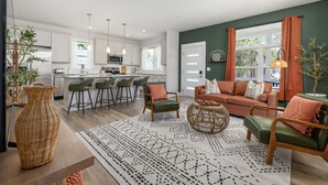 A modern living room with a sofa, chairs, and a coffee table. There is a kitchen in the background with white cabinets and a dining area with bar stools. - Brand New 4bd - Modern - Prime Location - Spacious (Raleigh)