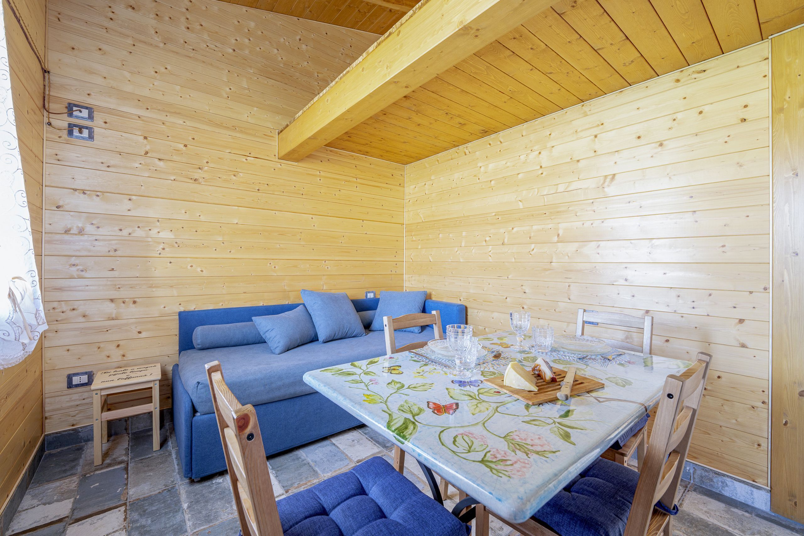 L'intérieur d'un chalet en bois, avec un canapé bleu, une table à manger recouverte d'une nappe à motifs floraux et un petit tabouret en bois.