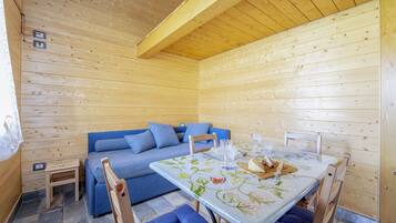 L'intérieur d'un chalet en bois, avec un canapé bleu, une table à manger recouverte d'une nappe à motifs floraux et un petit tabouret en bois.
