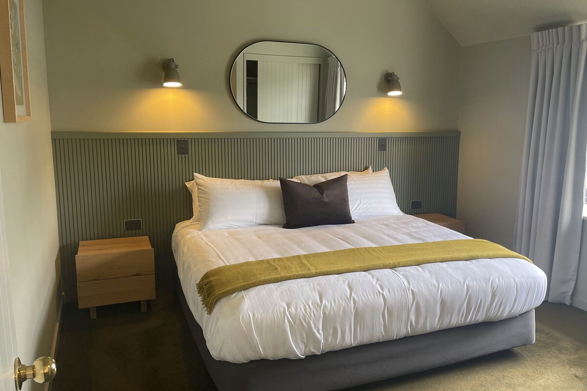 Een hotelkamer met een groot bed, twee bedlampen, een spiegel boven het bed, een nachtkastje en een raam met gordijnen.
