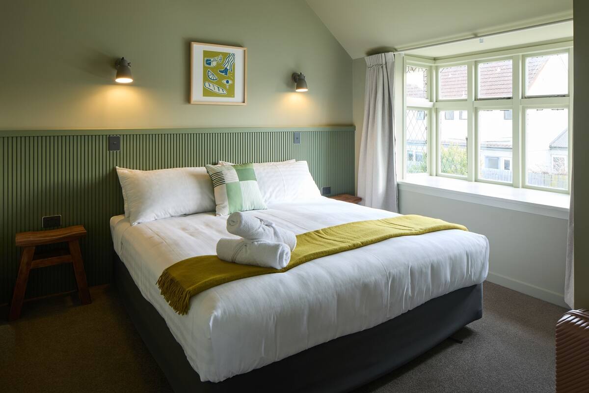 Een netjes opgemaakt bed met witte lakens en een groene plaid, een nachtkastje met een lamp, en een raam met gordijnen.