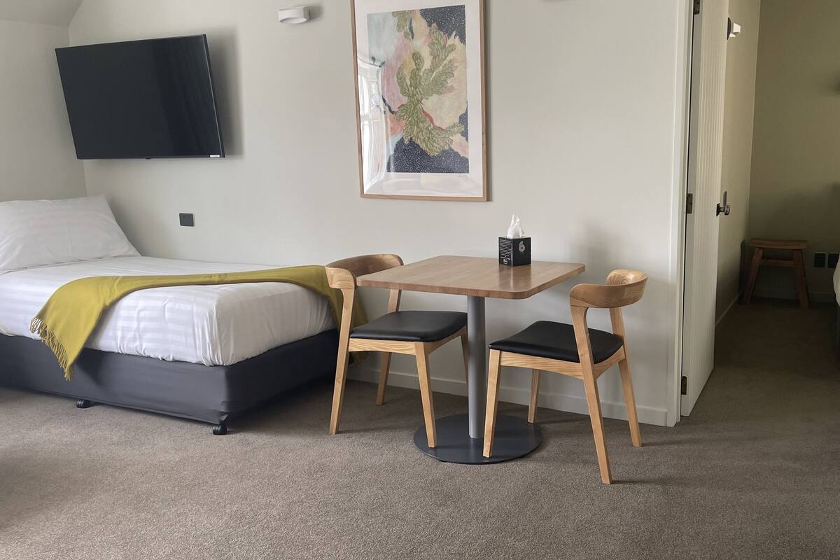 Een hotelkamer met een bed, een bureau met twee stoelen, een flatscreen televisie, een schilderij en een thermostaat.