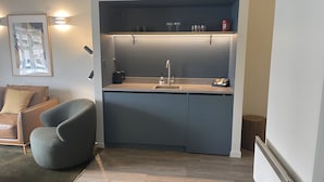 Una cocina compacta con lavamanos, encimera y armarios. En la zona de estar contigua hay una silla y una pequeña mesa.