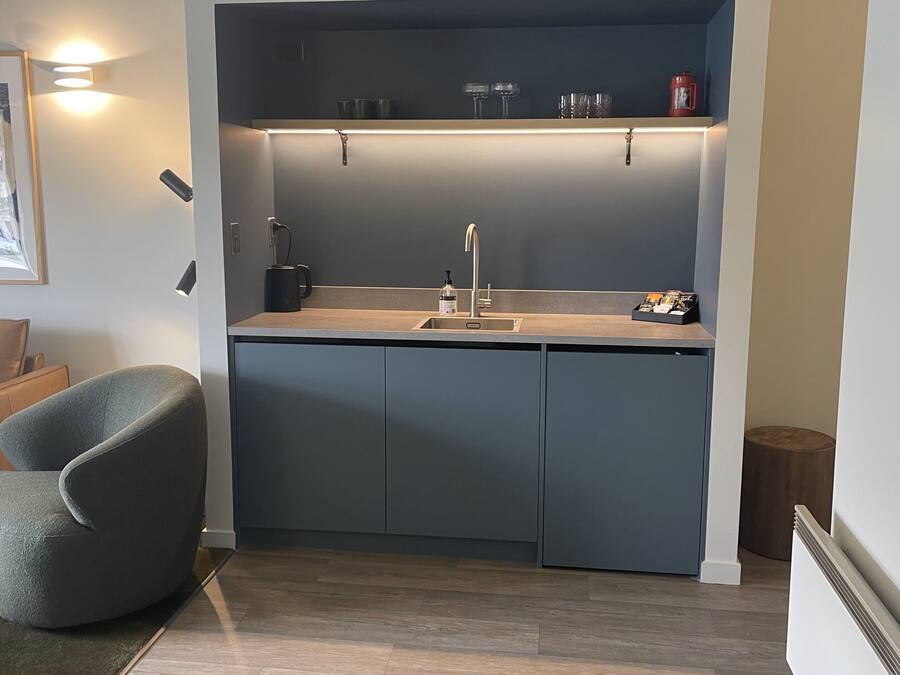 A compact kitchen with a sink, countertop, and cabinets. There is a chair and a small table in the adjacent living area.