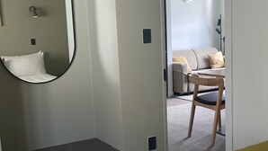 Un dormitorio con una cama, un espejo circular, un estante pequeño y una puerta que da a otra habitación.