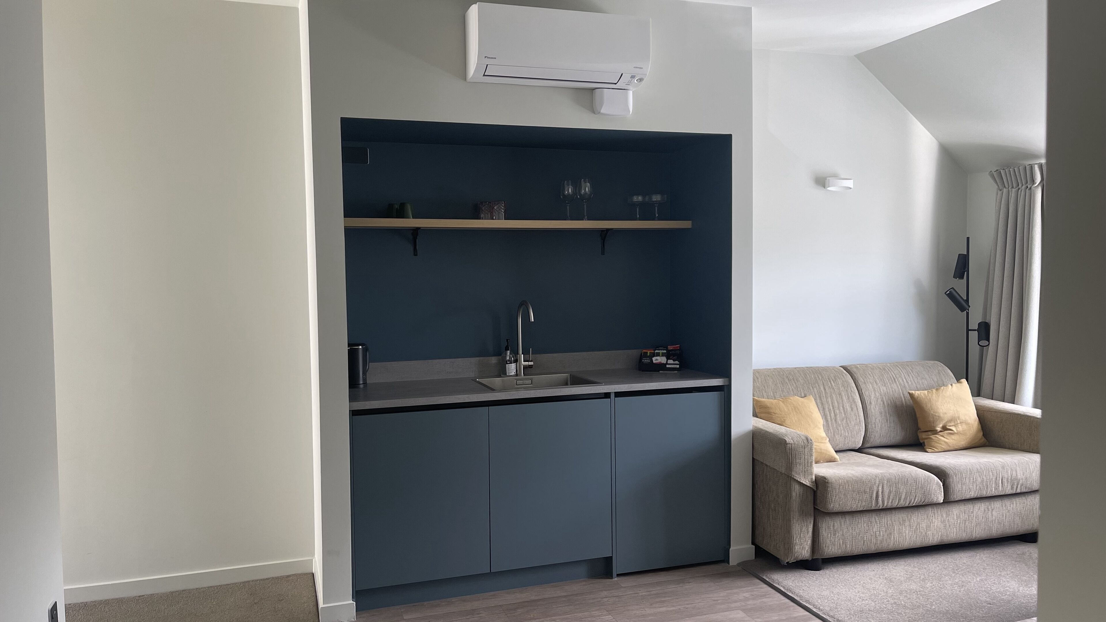 A compact kitchen with a built-in sink and a dark blue upper shelf, adjacent to a grey sofa with a cushion.