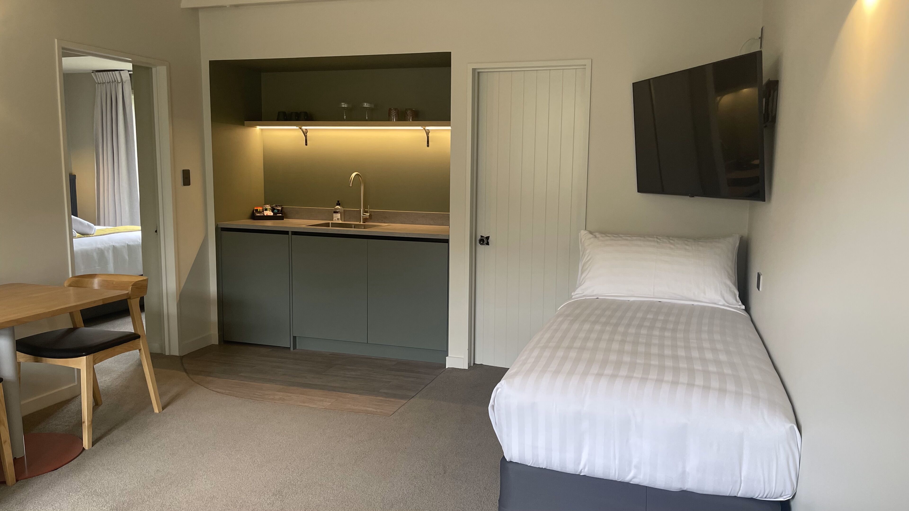 Une pièce de taille modeste comprenant un lit, un bureau avec une chaise, une petite kitchenette et un téléviseur fixé au mur.