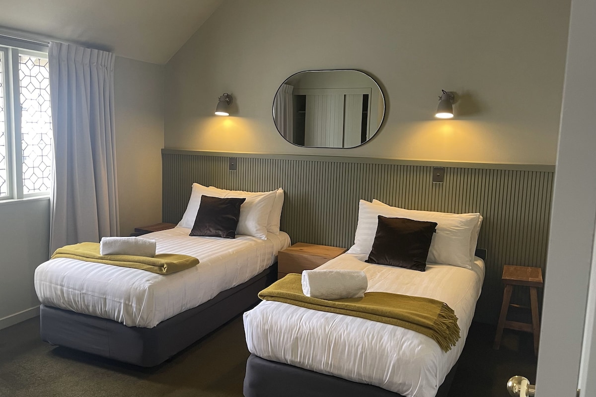 Une chambre d’hôtel avec deux lits simples, chacun équipé d’un oreiller, d’une couverture et d’une table de chevet. La chambre dispose d’une fenêtre avec des rideaux, d’un miroir fixé au mur et de deux appliques murales qui fournissent de l’éclairage.